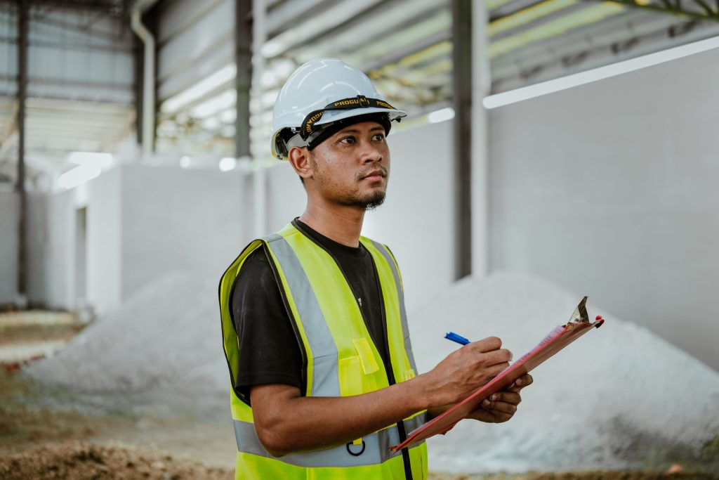 Site supervisor in a hard hat and high-visibility vest holding a clipboard in a factory in Banting, Selangor