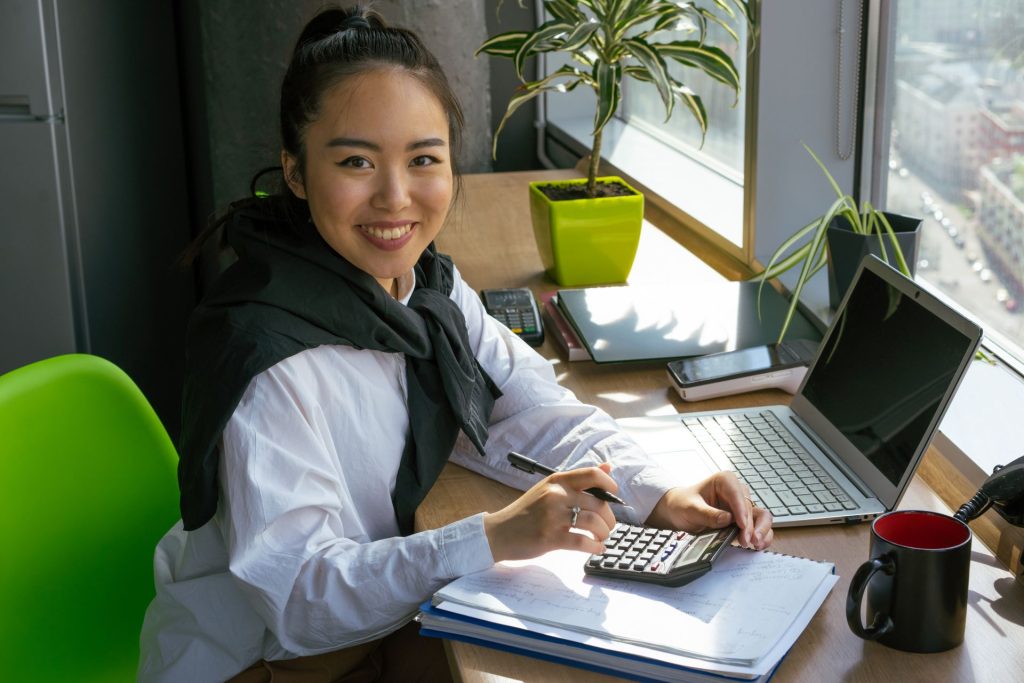 Smiling woman using a calculator beside a laptop and paperwork